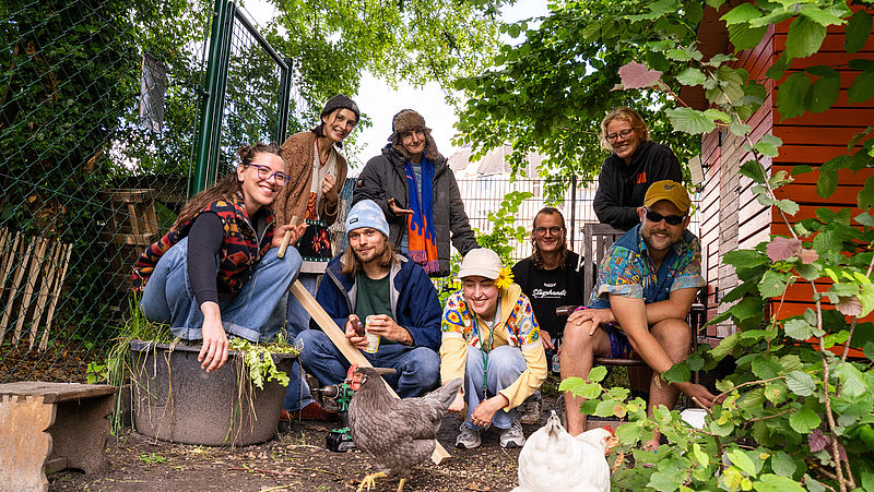 Menschen stehen/ sitzen draußen im Hühnerstall. Im Vordergrund sind zwei Hühner zu sehen, die gefüttert werden