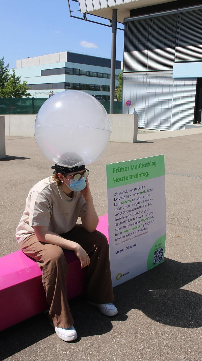 Person trägt eine mit Trockennebel gefüllte Kugel auf dem Kopf. Hinter der Person wird auf einem Plakat das Symptom Brainfog bei ME/CFS erklärt.