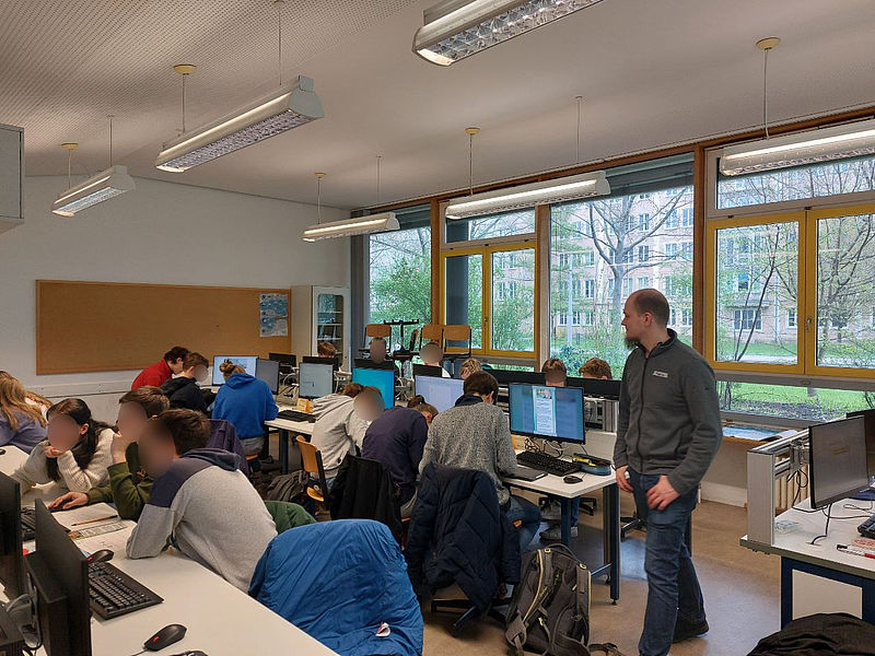 Eine Schulklasse sitzt in Gruppen an Computern. Die Gruppen analysieren Nachrichten und diskutieren ihre Glaubhaftigkeit. Ein Teamer läuft durch die Reihen, um Hilfestellungen zu geben.