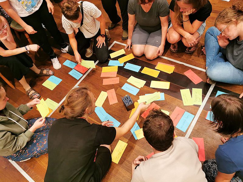 Jugendliche sitzen eng beieinander in einem Quadrat. Die Teilnehmenden halten sich gegenseitig an den Händen oder haken sich ein.