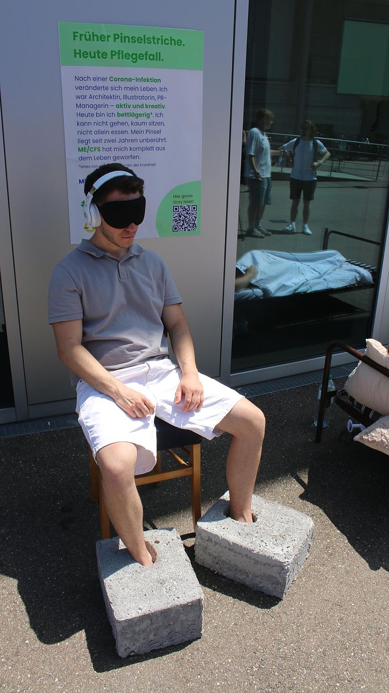 Person sitzt mit "Beton an den Füßen", Sonnenbrille und Gehörschutz auf einem Stuhl. Hinter der Person wird auf einem Plakat das Symptom Muskelschwäche bei ME/CFS erklärt.