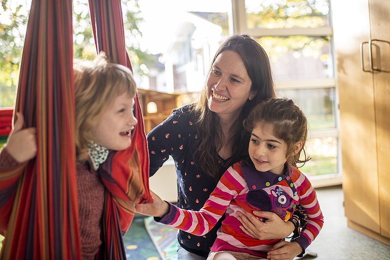 Eine Frau sitzt mit einem Mädchen auf dem Schoß auf dem Boden und lacht. Ein anderes Kind sitzt in einer Schaukel daneben und lacht.