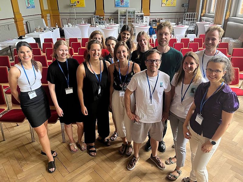 Eine Gruppe junger Menschen steht in einem Raum vor Stühlen und schaut in die Kamera