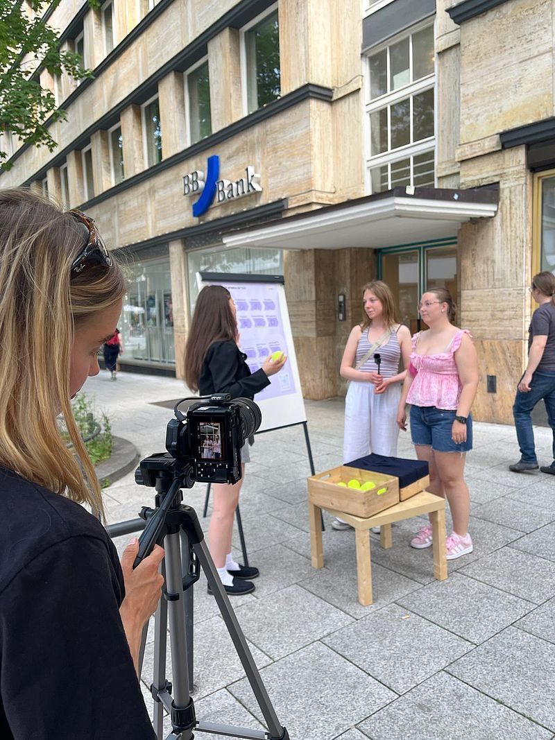 Person sitzt mit "Beton an den Füßen", Sonnenbrille und Gehörschutz auf einem Stuhl. Hinter der Person wird auf einem Plakat das Symptom Muskelschwäche bei ME/CFS erklärt.
