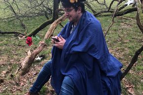 Ein Mann sitzt draußen bei einem Baum und hat einen blauen Umhang um.